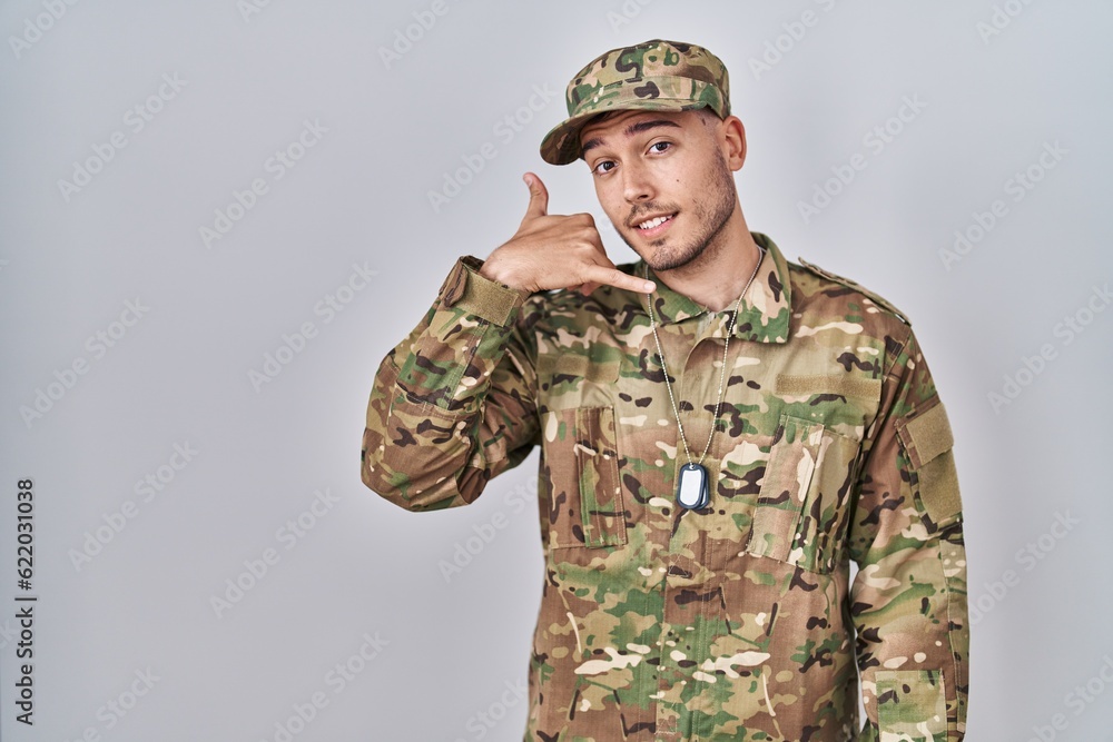Young hispanic man wearing camouflage army uniform smiling doing phone gesture with hand and fingers like talking on the telephone. communicating concepts. Young hispanic man wearing camouflage army uniform smiling doing phone gesture with hand and fingers like talking on the telephone. communicating concepts.