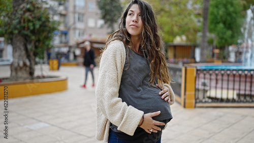 Young pregnant woman standing with serious expression touching belly at park