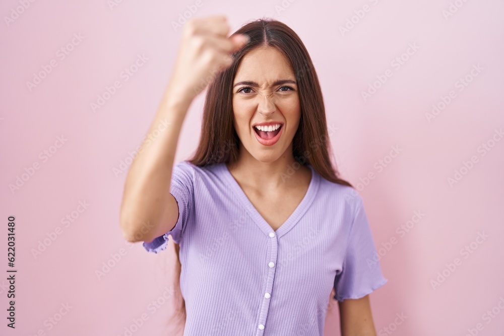 Young hispanic woman with long hair standing over pink background angry and mad raising fist frustrated and furious while shouting with anger. rage and aggressive concept. Young hispanic woman with long hair standing over pink background angry and mad raising fist frustrated and furious while shouting with anger. rage and aggressive concept.