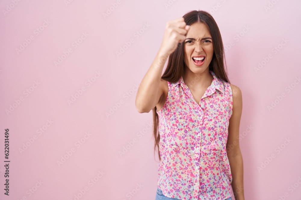Young hispanic woman with long hair standing over pink background angry and mad raising fist frustrated and furious while shouting with anger. rage and aggressive concept. Young hispanic woman with long hair standing over pink background angry and mad raising fist frustrated and furious while shouting with anger. rage and aggressive concept.