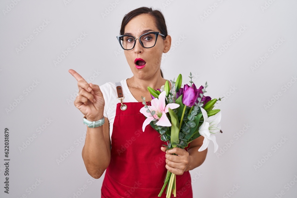Middle age brunette woman wearing apron working at florist shop holding bouquet surprised pointing with finger to the side, open mouth amazed expression. Middle age brunette woman wearing apron working at florist shop holding bouquet surprised pointing with finger to the side, open mouth amazed expression.