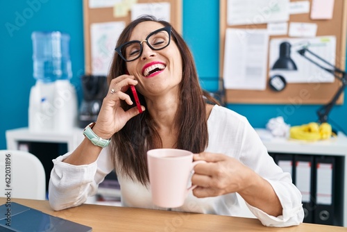 Young beautiful hispanic woman business worker talking on smartphone drinking coffee at office