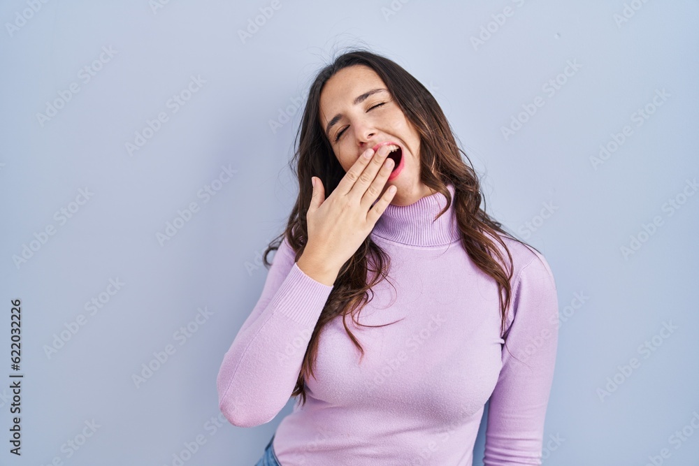 Young brunette woman standing over blue background bored yawning tired covering mouth with hand. restless and sleepiness. Young brunette woman standing over blue background bored yawning tired covering mouth with hand. restless and sleepiness.