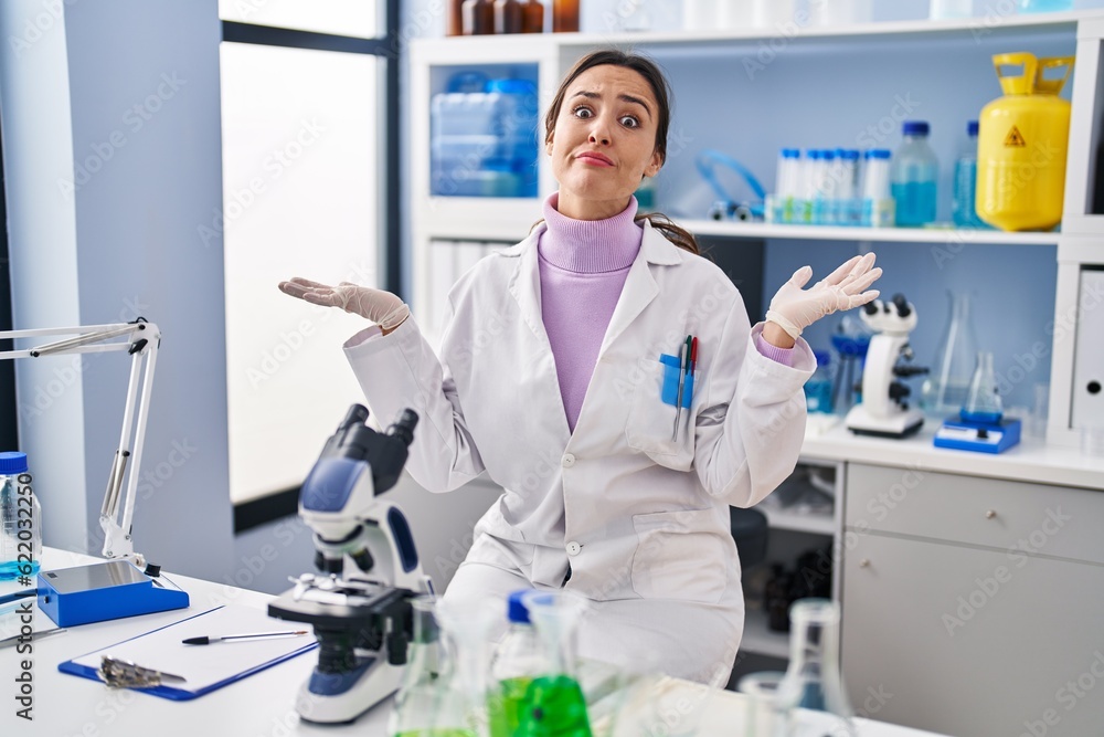 Young brunette woman working at scientist laboratory clueless and confused expression with arms and hands raised. doubt concept. Young brunette woman working at scientist laboratory clueless and confused expression with arms and hands raised. doubt concept.