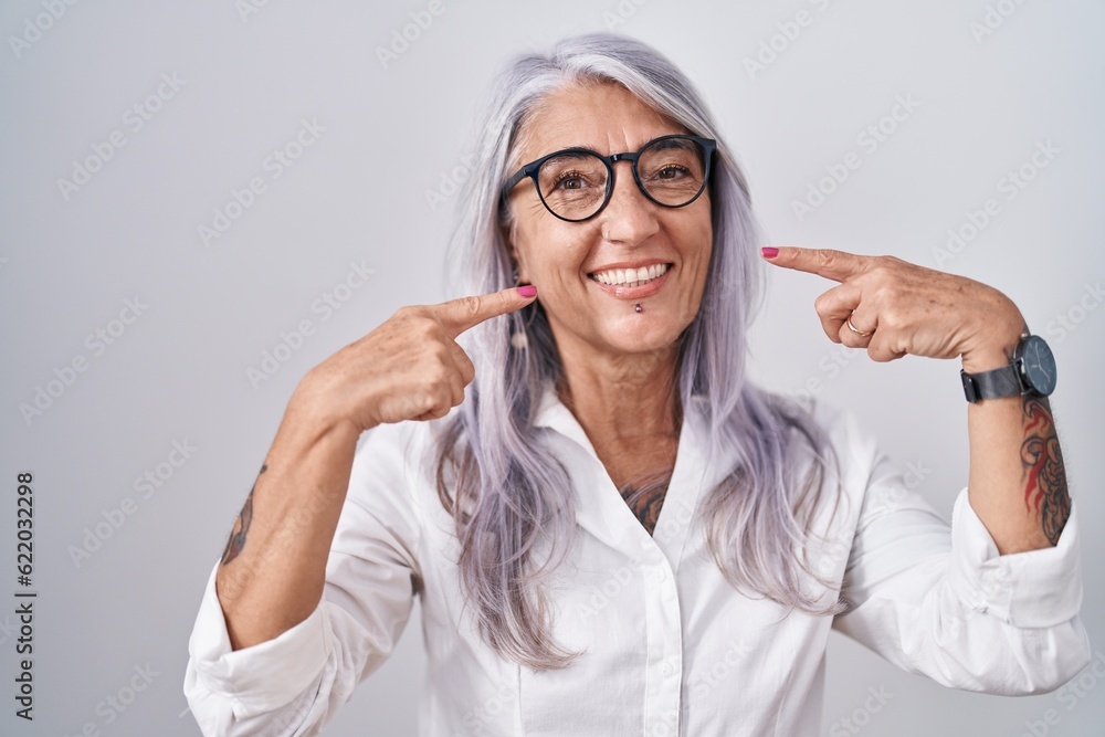 Middle age woman with tattoos wearing glasses standing over white background smiling cheerful showing and pointing with fingers teeth and mouth. dental health concept. Middle age woman with tattoos wearing glasses standing over white background smiling cheerful showing and pointing with fingers teeth and mouth. dental health concept.