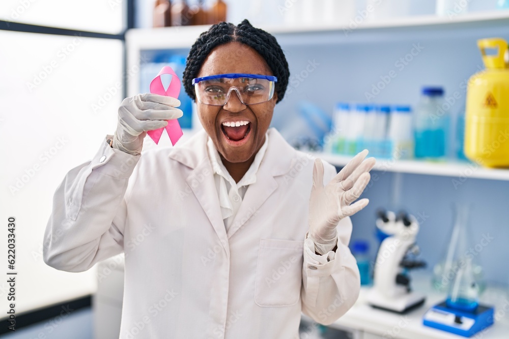 African american woman working at scientist laboratory holding pink ribbon celebrating victory with happy smile and winner expression with raised hands African american woman working at scientist laboratory holding pink ribbon celebrating victory with happy smile and winner expression with raised hands