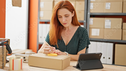 Young redhead woman ecommerce business worker writing on reminder paper using touchpad at office