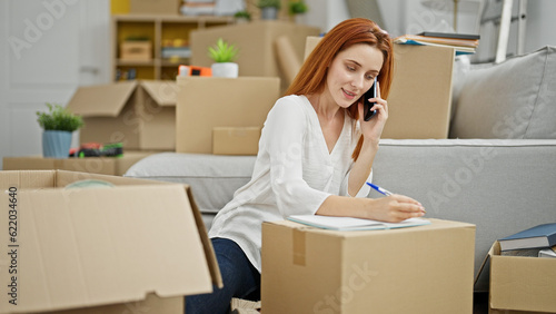 Young redhead woman talking on smartphone writing on notebook at new home