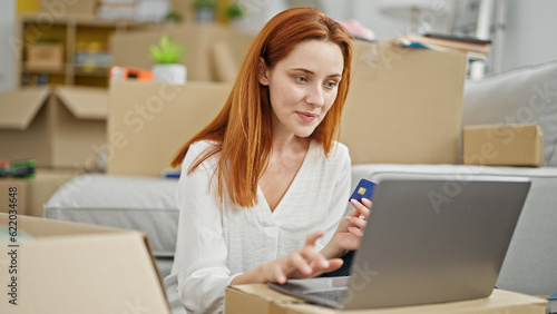 Young redhead woman shopping with laptop and credit card sitting on floor at new home