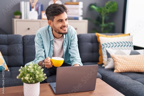 Young hispanic man using laptop drinking coffee at home