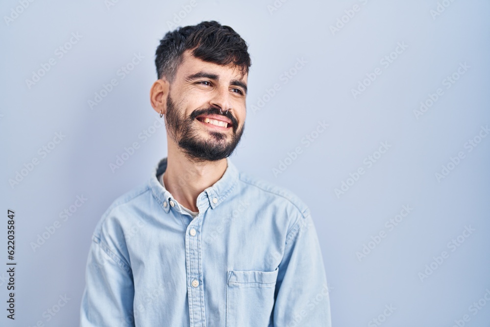 Young hispanic man with beard standing over blue background looking away to side with smile on face, natural expression. laughing confident. Young hispanic man with beard standing over blue background looking away to side with smile on face, natural expression. laughing confident.