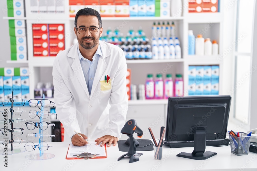 Young hispanic man pharmacist smiling confident writing on document at pharmacy