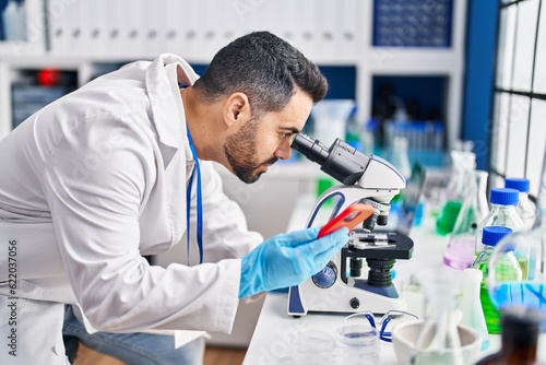 Young hispanic man scientist using microscope and smartphone at laboratory