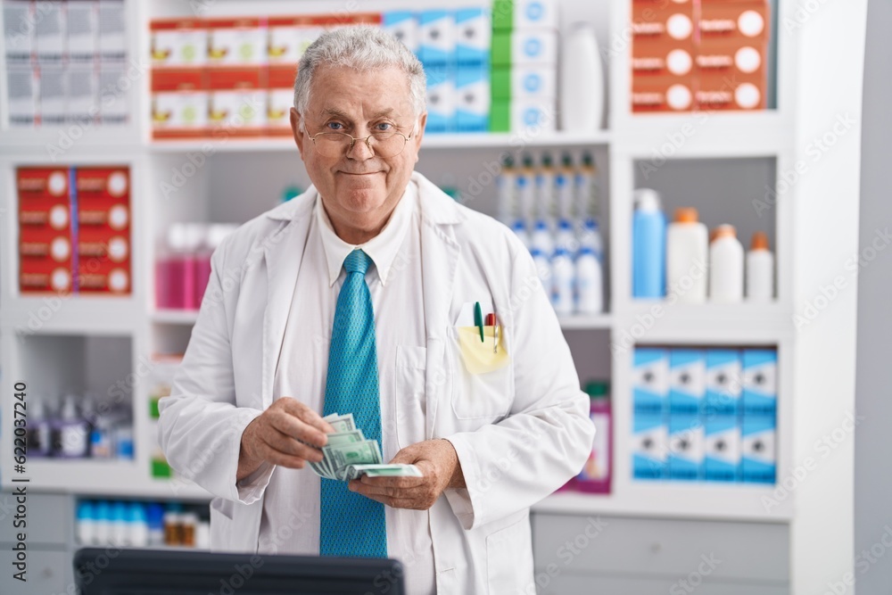 Middle age grey-haired man pharmacist smiling confident counting dollars at pharmacy