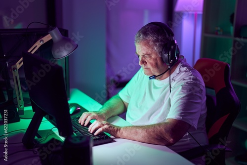 Middle age grey-haired man streamer playing video game using computer at gaming room