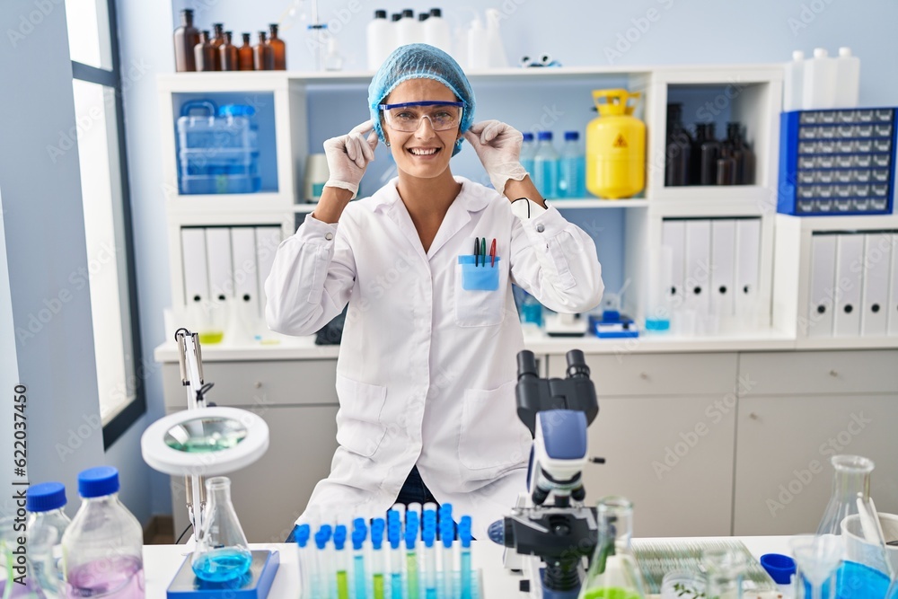 Brunette woman working at scientist laboratory smiling pulling ears with fingers, funny gesture. audition problem Brunette woman working at scientist laboratory smiling pulling ears with fingers, funny gesture. audition problem