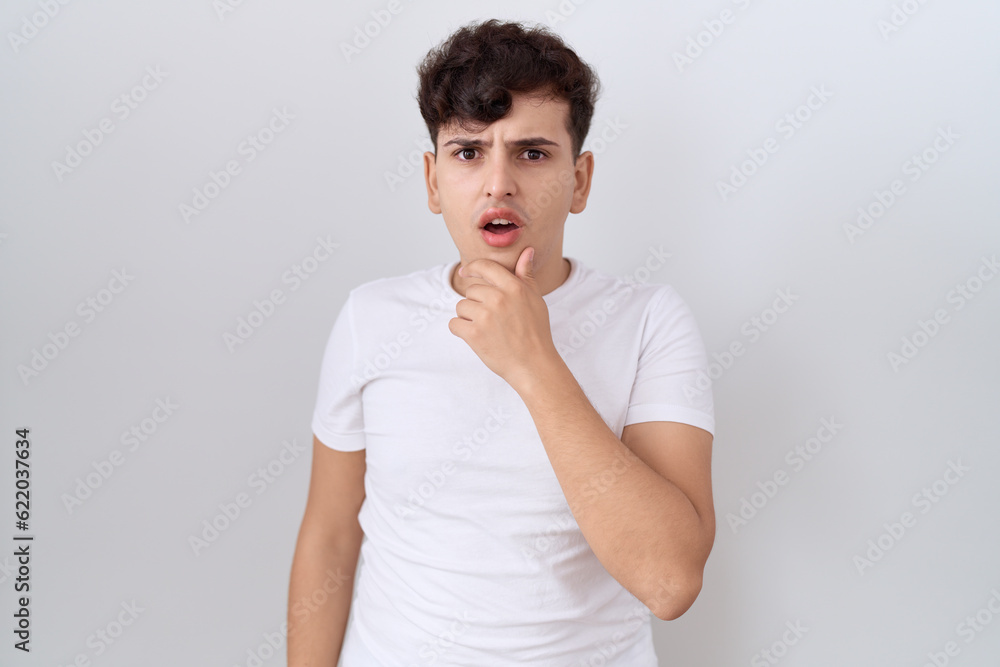Young non binary man wearing casual white t shirt looking fascinated with disbelief, surprise and amazed expression with hands on chin Young non binary man wearing casual white t shirt looking fascinated with disbelief, surprise and amazed expression with hands on chin