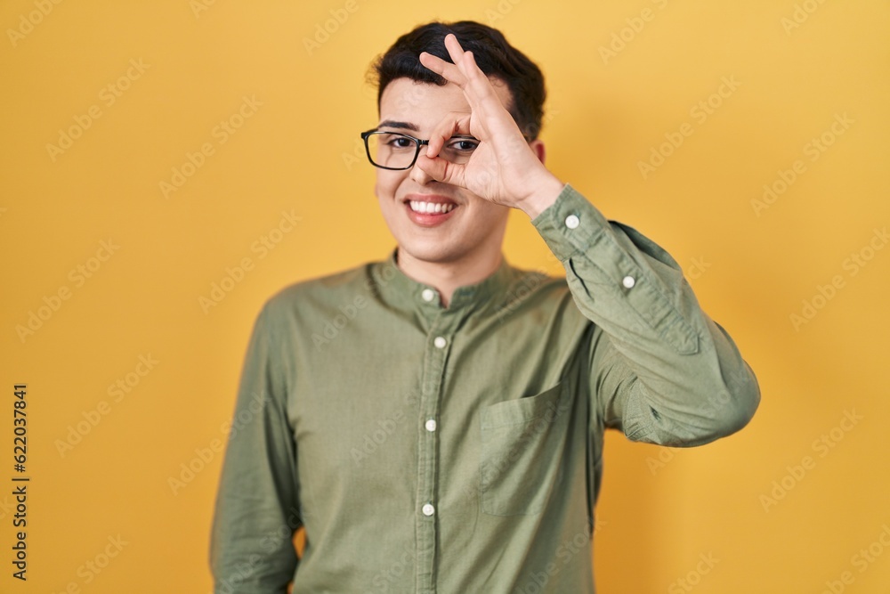 Non binary person standing over yellow background doing ok gesture with hand smiling, eye looking through fingers with happy face. Non binary person standing over yellow background doing ok gesture with hand smiling, eye looking through fingers with happy face.