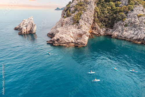 Photography Aerial view of a group or team with paddles swims on a SUP boards on the sea near scenic rocks