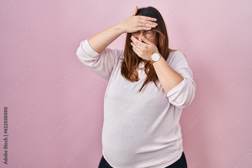 Pregnant woman standing over pink background covering eyes and mouth with hands, surprised and shocked. hiding emotion Pregnant woman standing over pink background covering eyes and mouth with hands, surprised and shocked. hiding emotion
