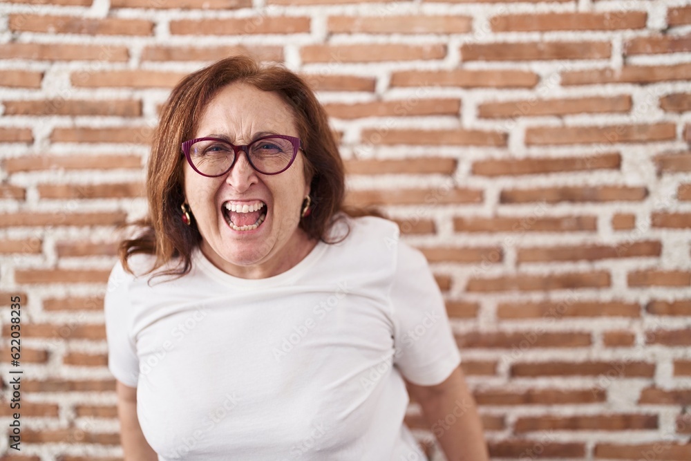 Senior woman with glasses standing over bricks wall angry and mad screaming frustrated and furious, shouting with anger. rage and aggressive concept. Senior woman with glasses standing over bricks wall angry and mad screaming frustrated and furious, shouting with anger. rage and aggressive concept.