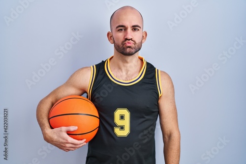 Young bald man with beard wearing basketball uniform holding ball looking at the camera blowing a kiss on air being lovely and sexy. love expression.