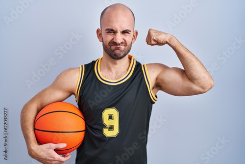 Young bald man with beard wearing basketball uniform holding ball strong person showing arm muscle, confident and proud of power