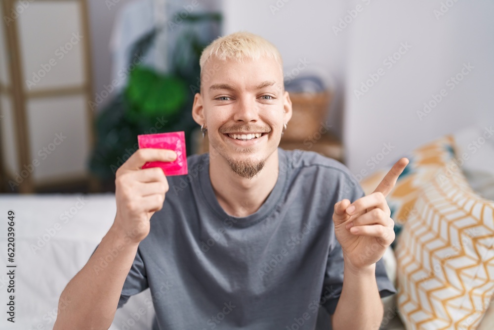 Young caucasian man holding condom sitting on bed smiling happy pointing with hand and finger to the side Young caucasian man holding condom sitting on bed smiling happy pointing with hand and finger to the side