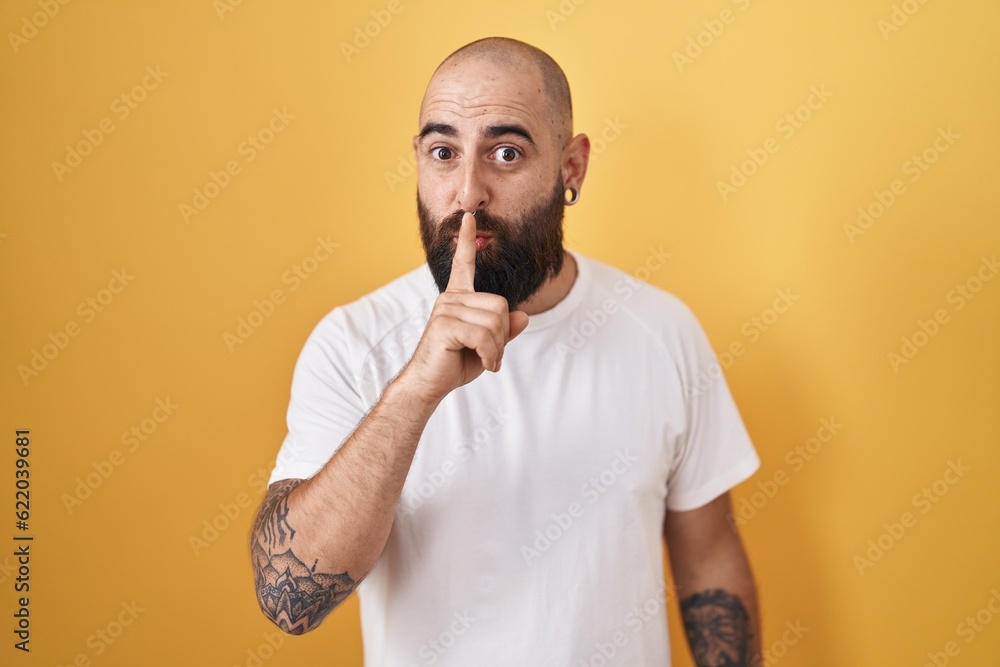 Young hispanic man with beard and tattoos standing over yellow background asking to be quiet with finger on lips. silence and secret concept. Young hispanic man with beard and tattoos standing over yellow background asking to be quiet with finger on lips. silence and secret concept.
