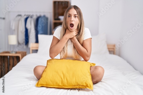 Young blonde woman sitting on the bed with pillow at home shouting and suffocate because painful strangle. health problem. asphyxiate and suicide concept.