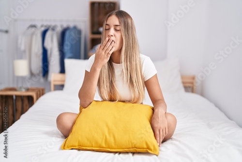 Young blonde woman sitting on the bed with pillow at home bored yawning tired covering mouth with hand. restless and sleepiness.