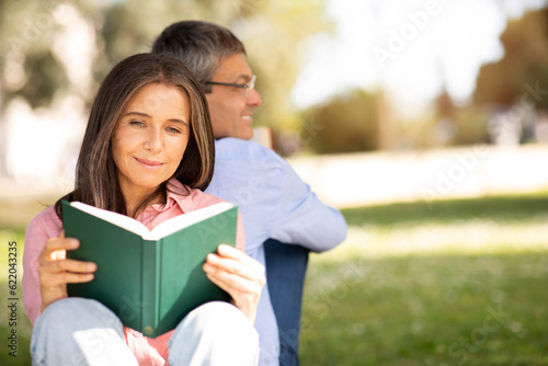Beautiful Mature Woman Reading Book While Relaxing In Park With Husband
