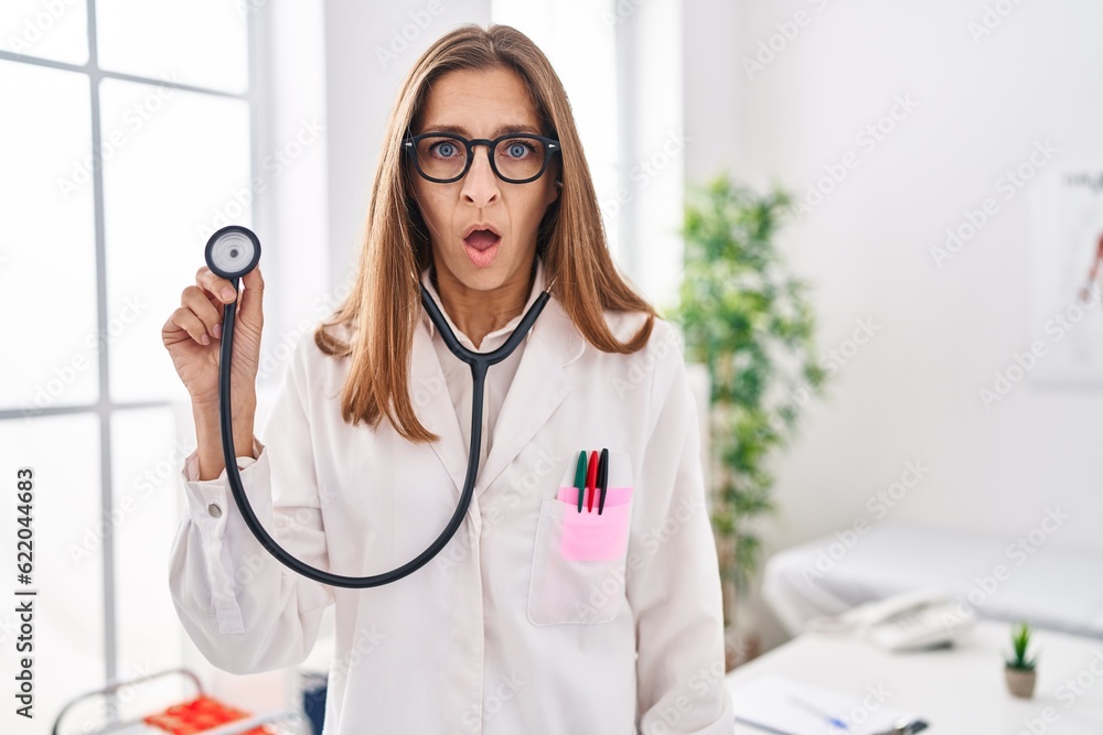 Young woman wearing doctor uniform holding stethoscope scared and amazed with open mouth for surprise, disbelief face Young woman wearing doctor uniform holding stethoscope scared and amazed with open mouth for surprise, disbelief face