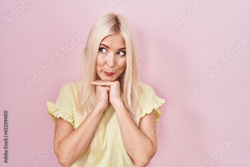 Caucasian woman standing over pink background laughing nervous and excited with hands on chin looking to the side