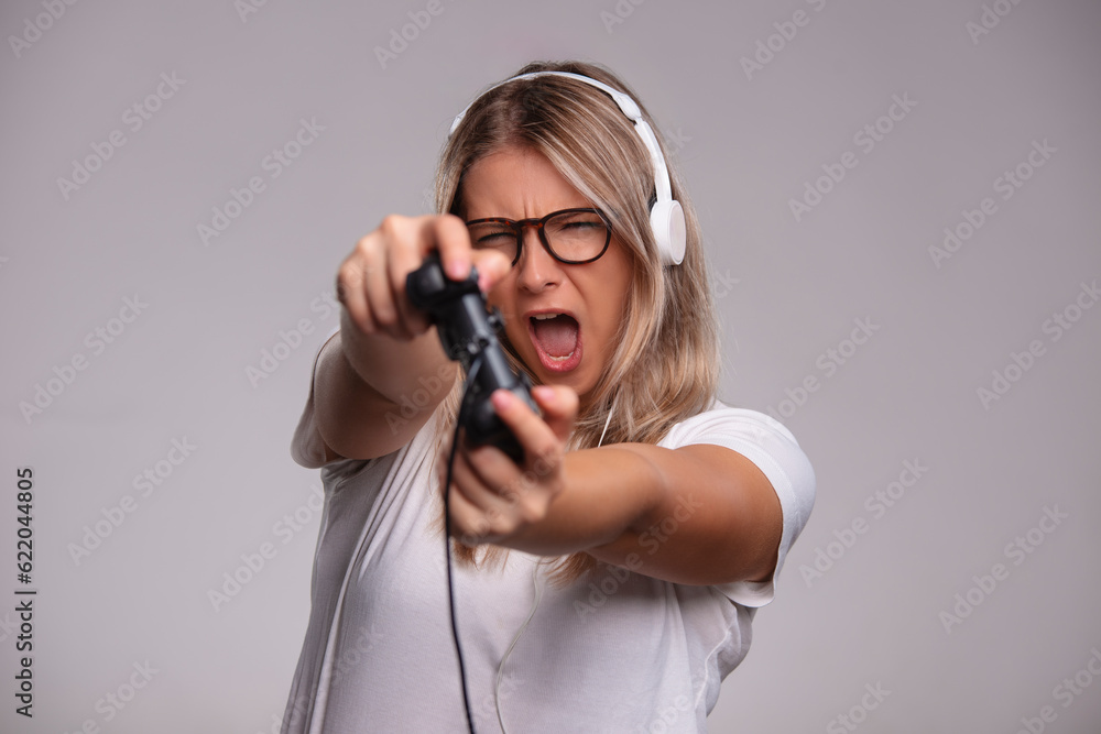Young beautiful gamer woman playing video game and using joystick and headphones on grey background Young beautiful gamer woman playing video game and using joystick and headphones on grey background