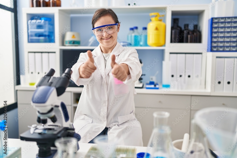 Hispanic girl with down syndrome working at scientist laboratory success sign doing positive gesture with hand, thumbs up smiling and happy. cheerful expression and winner gesture. Hispanic girl with down syndrome working at scientist laboratory success sign doing positive gesture with hand, thumbs up smiling and happy. cheerful expression and winner gesture.
