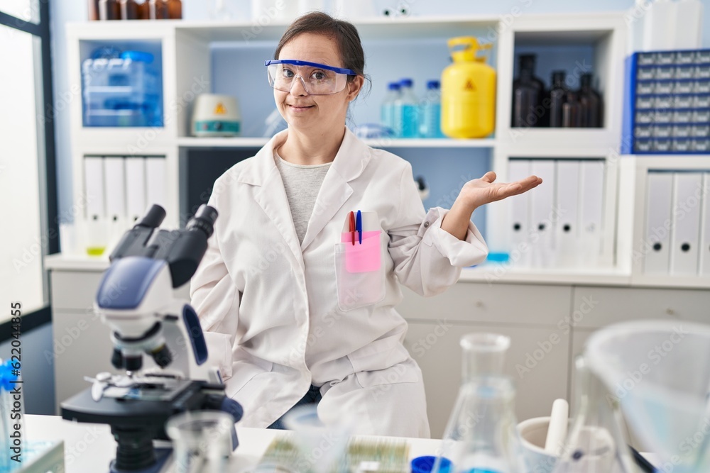 Hispanic girl with down syndrome working at scientist laboratory smiling cheerful presenting and pointing with palm of hand looking at the camera. Hispanic girl with down syndrome working at scientist laboratory smiling cheerful presenting and pointing with palm of hand looking at the camera.