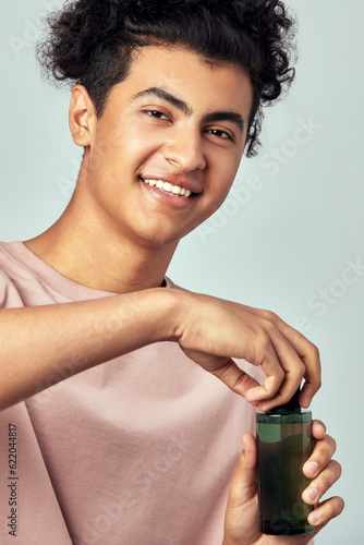 Portrait of young handsome smiling guy with a bottle of facial lotion in his hands. Swarthy man with black curly hair practice skincare routine to maintain his youthful and healthy appearance.
