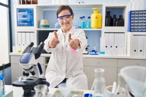 Hispanic girl with down syndrome working at scientist laboratory success sign doing positive gesture with hand  thumbs up smiling and happy. cheerful expression and winner gesture.