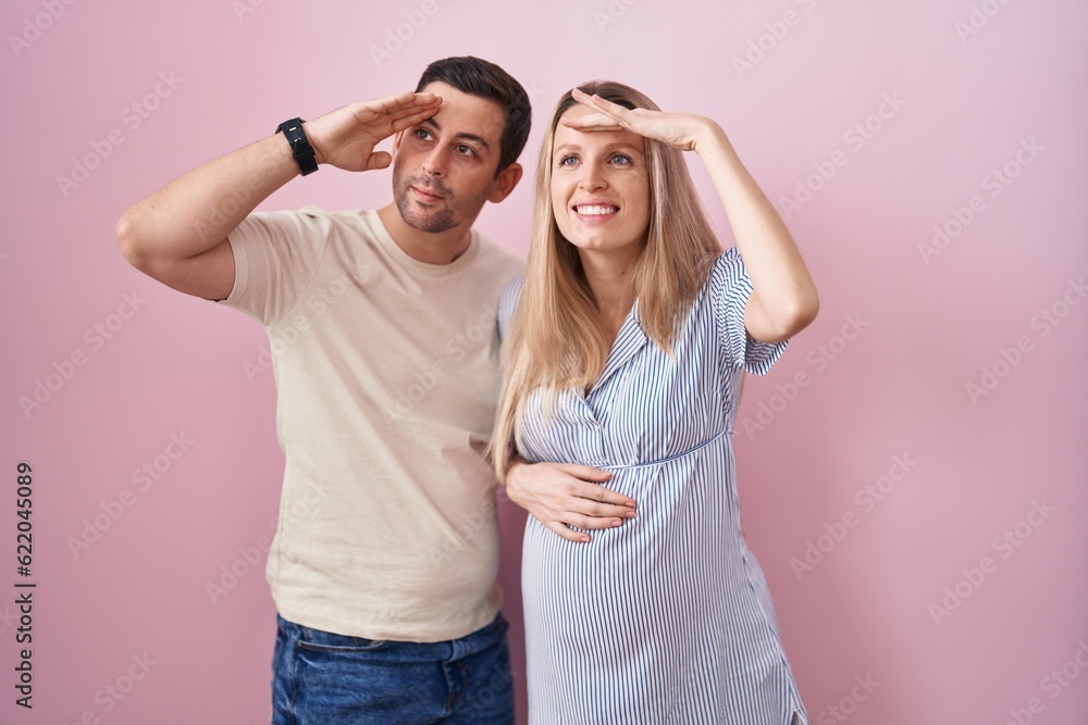 Young couple expecting a baby standing over pink background very happy and smiling looking far away with hand over head. searching concept. Young couple expecting a baby standing over pink background very happy and smiling looking far away with hand over head. searching concept.