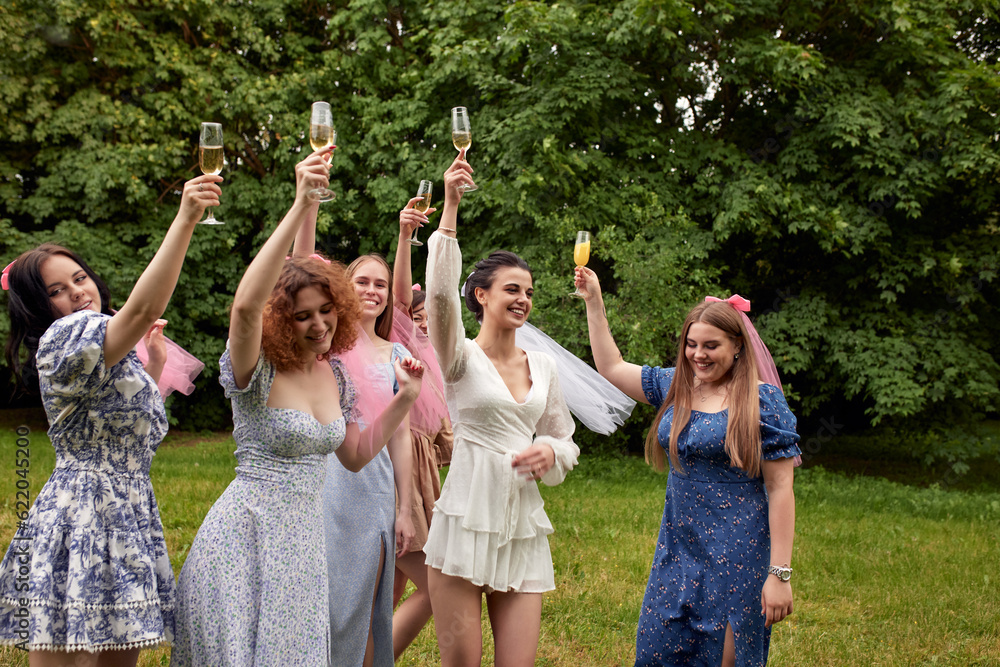 Obraz premium Happy charming bride in white dress with white veil and bridesmaids with pink veils raise champagne glasses and laugh merrily. Cheerful bachelorette party in the summer park.