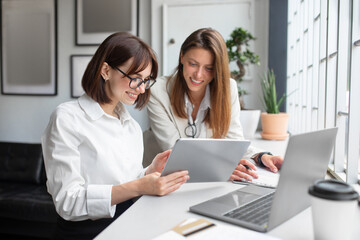 Two lady colleagues discussing new project, businesswomen using digital tablet together in office