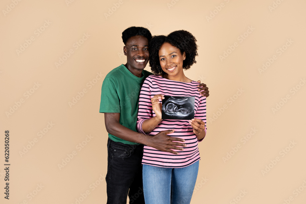 Fototapeta premium Smiling millennial african american husband hugging woman with big belly, show ultrasound scan