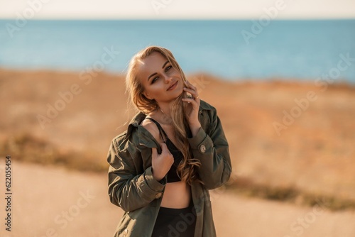 Portrait blonde sea cape. A calm young blonde in an unbuttoned khaki raincoat stands on the seashore  under the raincoat there is a black skirt and top