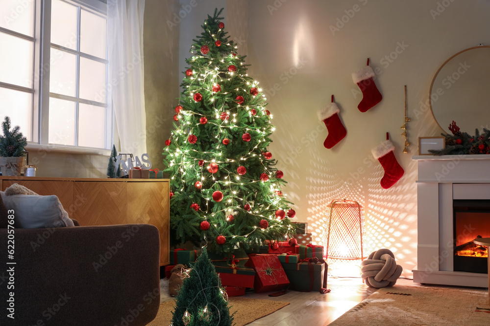 Christmas tree with gifts, glowing lights and fireplace in dark living room