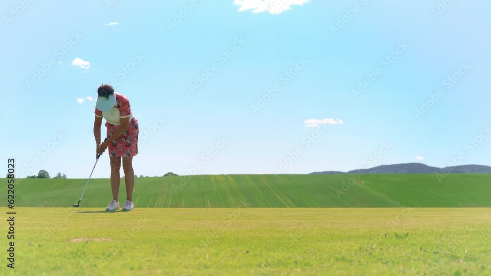 Mature woman playing golf. Golfer hitting golf shot and hammering a ball into the golf hole. Beautiful sunny landscape, with green hills and blue sky. Mature woman playing golf. Golfer hitting golf shot and hammering a ball into the golf hole. Beautiful sunny landscape, with green hills and blue sky.