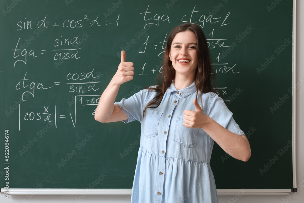 Female Math teacher showing thumbs-up near chalkboard in classroom Female Math teacher showing thumbs-up near chalkboard in classroom