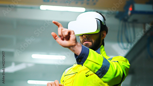 Close up of Caucasian male young engineer in headset and yellow uniform standing in factory and having virtual reality experience. Engineering concept. Indoors. Man in VR goggles moving hands in air.