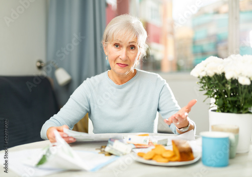 Positive aged woman calculating running costs of household chores sitting at home table with money and reading papers.