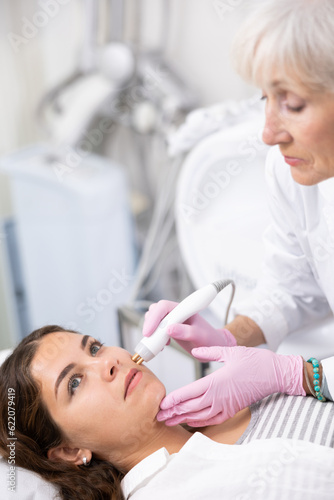 Positive young woman having radiolifting of her face by means of apparatus for aesthetic procedures used by specialist